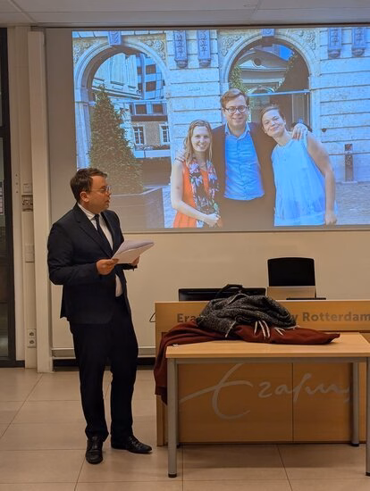 Man in suit holding papers at classroom lectern, projected photo of four smiling adults.