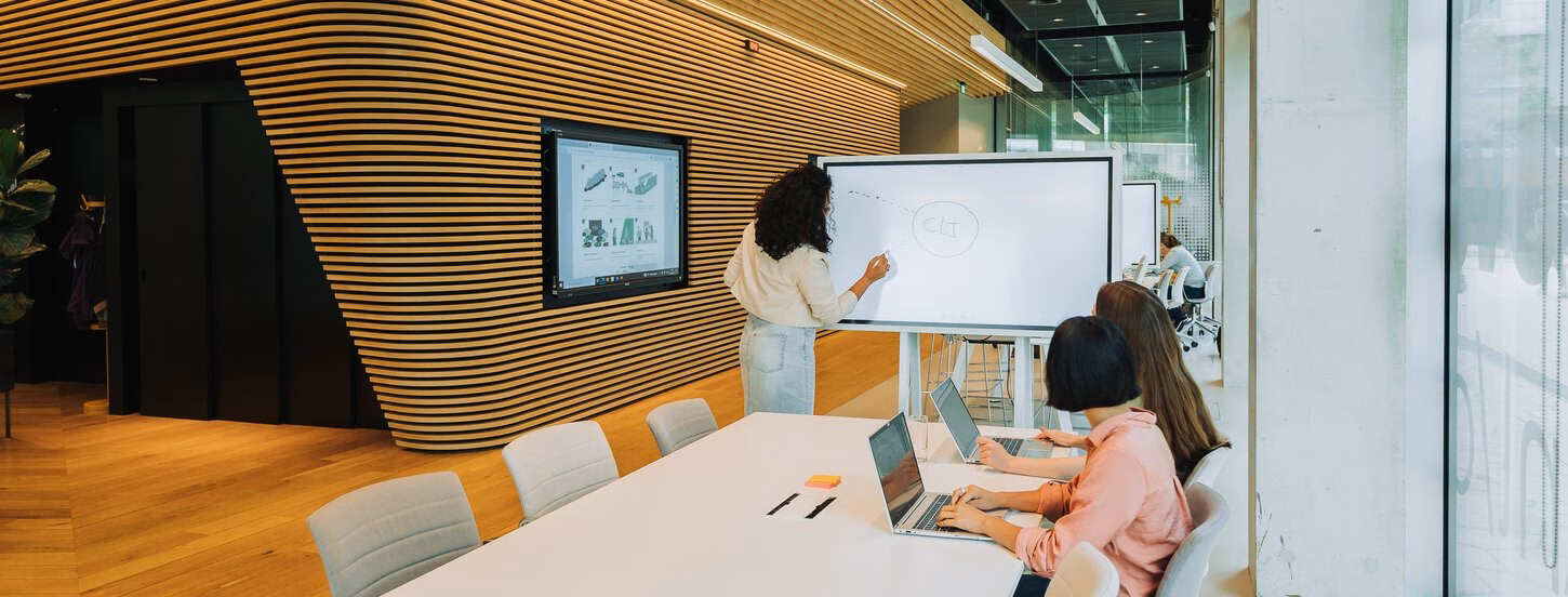 Two people at a meeting table, watching someone draw on a smart screen.