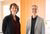 Photograph of two businesspeople standing indoors by wooden panels, faces blurred.