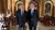 Two smiling men in suits standing in an ornate hall with arched doorways and wooden chairs.