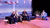 Photograph showing four panelists seated on stage under purple lighting.