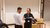 Woman in glasses handing an item to a man in a white shirt beside a classroom lectern