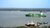 Green container ship crossing a wide river with tugboats near the riverbank under cloudy sky