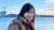 Woman in a brown coat smiling by a waterfront with factory chimneys in the background