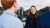 Two people facing a blurred person in a black coat by a lakeside on a cloudy day