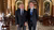 Two smiling men in suits standing in an ornate hall with arched doorways and wooden chairs.