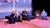 Photograph showing four panelists seated on stage under purple lighting.