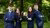 Four smiling young professionals in navy suits standing back-to-back with crossed arms in leafy park