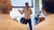 Man gesturing while presenting by projector screen to seated audience in sunlit classroom.