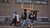 Photograph of group carrying assorted furniture along brick sidewalk, including a chair overhead