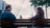 Een man en een vrouw in gesprek op een bankje bij de waterkant, met op de achtergrond de skyline van Rotterdam bij bewolkt weer.