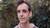 Photograph of a man in a pink shirt gazing off camera against blurred green foliage.