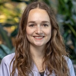 Woman with long wavy brown hair wearing lavender shirt in leafy setting